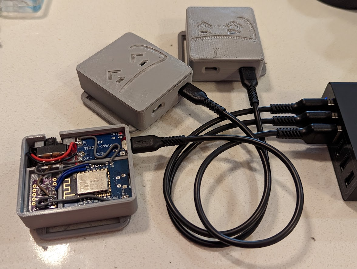 three grey slimeVR body trackers with one case open, exposing the circuits within. All three are plugged into a usb hub with black attached to black cables.